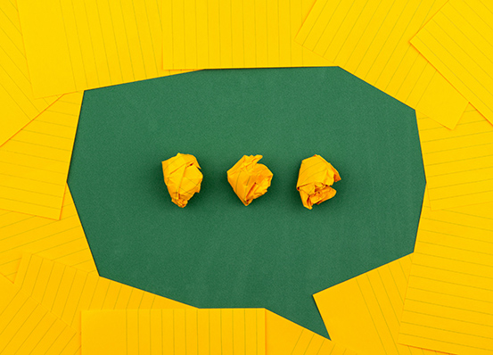 A flat lay of yellow papers forming a dialogue box with crumbled yellow papers in the box in the shape of an ellipsis. 